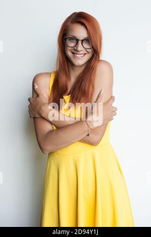 Rothaarige Frau mit Brille umarmt sich trägt ein Gelbes Kleid auf einer weißen Studiowand lächelt der Kamera zu Stockfoto