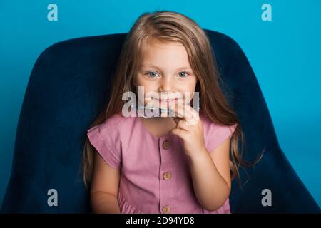 Charming blonde Mädchen lächelt in einem Sessel abheben Ihre Brille auf einer blauen Studiowand Stockfoto