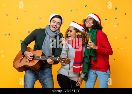 Glücklich drei multirassische Freunde singen zusammen in Weihnachtsfeier Auf gelbem Studiohintergrund mit Konfetti Stockfoto