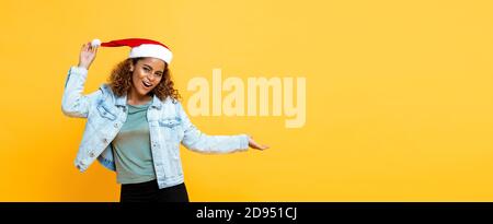 Fun Taille bis Porträt von fröhlichen afroamerikanischen Frau tragen Weihnachtsmütze mit offener Palmgeste auf gelbem isoliertem Banner Hintergrund mit SPA kopieren Stockfoto