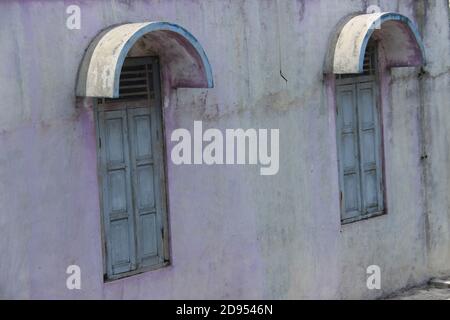 Altes Haus mit zwei blauen Holzfenstern und einer kaputten Wand. Altes Haus im ländlichen indonesien. Moosige Wandstruktur für den Hintergrund Stockfoto