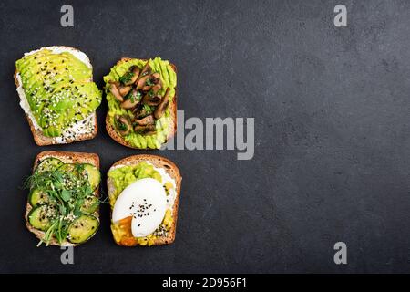 Set von Avocado Toasts mit verschiedenen Toppings. Vegetarischer Avocado-Toast mit Ei, gebratenen Pilzen, Gurken, Samen und Mikrogemüse auf schwarzem Beton Stockfoto