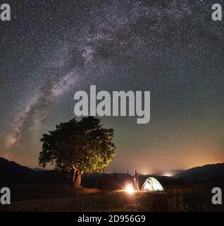 Weibliche Camper, die eine Pause auf Sommer Camping in den Bergen neben Lagerfeuer, glühenden touristischen Zelt und großen Baum. Junge Frau genießen Blick auf den Nachthimmel voller Sterne und Milchstraße. Astrofotografie Stockfoto