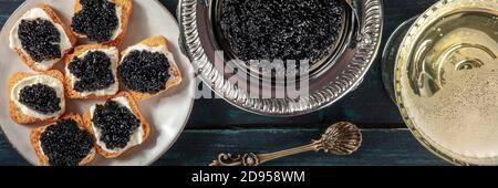 Schwarzes Kaviarpanorama, über dem Kopf flach liegend. Kaviar auf Toasts und in einer Vintage-Schüssel, mit einem Champagner-Coupé Stockfoto
