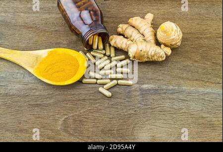 Kurkuma Kräuterkapseln in schwarzer Flasche, frische Kurkuma Wurzel und Kurkuma Pulver in Holzlöffel auf Holzgrund Stockfoto