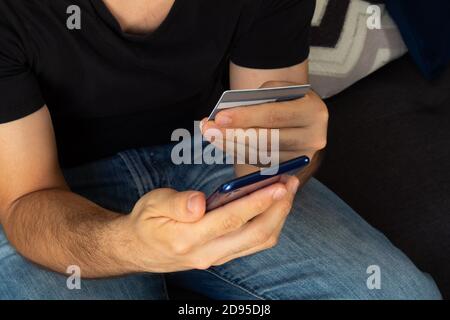 Nahaufnahme eines jungen hispanischen Mannes, der auf einem sitzt Sofa-Shopping online mit einem Smartphone und mit einem Kredit Karte Stockfoto