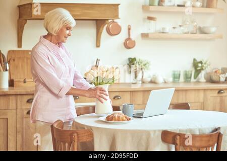 Glückliche ältere Frau, die morgens in der Küche eine Blumenvase auf den Tisch legt. Inländisches Lifestyle-Konzept Stockfoto