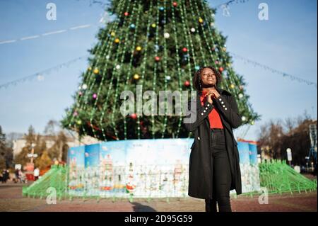 Porträt einer lockigen afrikanisch Frau tragen modischen schwarzen Mantel und roten Rollkragen posiert im Freien gegen Haupt-Neujahr Baum der Stadt. Stockfoto