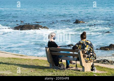 Zwei Frauen sitzen und plaudern auf einer Bank mit Blick auf das Meer Oder Meer während einer Pandemie tragen Gesichtsmasken und halten Ein Coronavirus mit sicherem Abstand Stockfoto