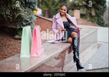 Junge stilvolle schöne afroamerikanische Frau in der Straße, tragen Mode-Outfit Mantel mit Einkaufstaschen und Handy an den Händen. Stockfoto