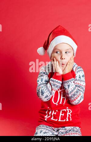 Neugieriges Kind in Weihnachten Pyjamas und Weihnachtsmann Hut schauen seitwärts in Überraschung und hält Wangen mit Händen, kopieren Raum Stockfoto