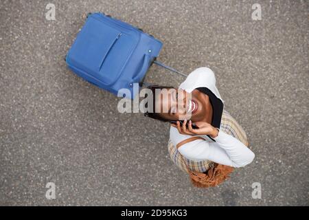 Portrait von oben von glücklicher afroamerikanischer Frau, die mit läuft Koffer und Handy Stockfoto