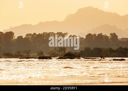 Laotische Fischer steuern ein Holzboot durch Stromschnellen auf dem Mekong Fluss in der Dämmerung, schwimmende Wald und Berge im Hintergrund. Champasak, Laos. Stockfoto