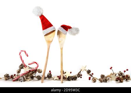 Zwei Kochlöffel aus Holz mit roten Weihnachtsmann-Hüten und einer weihnachtlichen Deko-Girlande, mit Zuckerstöcken, Zapfen, Sternen und künstlichen Beeren, isolieren Stockfoto