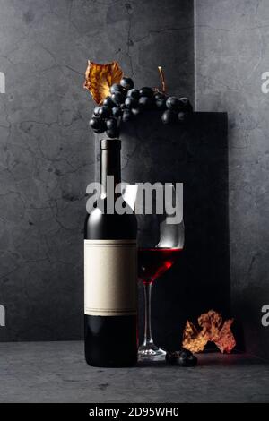 Rotwein und blaue Trauben mit ausgetrockneten Weinblättern auf einem schwarzen Steintisch. Auf einer Flasche altes leeres Etikett. Stockfoto