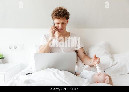 Glücklicher junger Vater, der mit seinem kleinen Sohn spielt, während er an einem Laptop arbeitet, im Bett liegt und auf dem Handy spricht Stockfoto