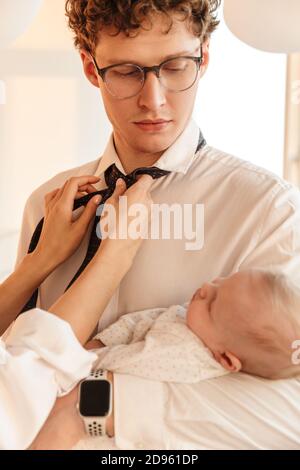 Frau Fixing Ehemänner Krawatte, während er hält ihr Baby Sohn, zu Hause stehen, immer bereit für die Arbeit Stockfoto