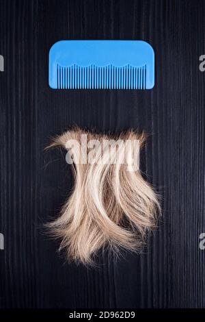 Blonde Haarsträhne, blauer Kunststoffkamm schwarzer Holzhintergrund Nahaufnahme, abgeschnittene natürliche blonde Haarlocken auf dunklem Holz, Haarbürste, Haarschnitt-Design Stockfoto