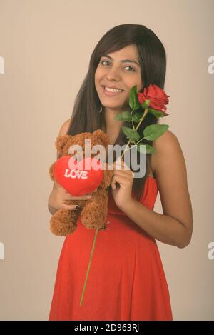 Junge glückliche persische Frau lächelt, während sie rote Rose und hält teddybär mit Herz und Liebe Zeichen bereit für Valentinstag Tag Stockfoto