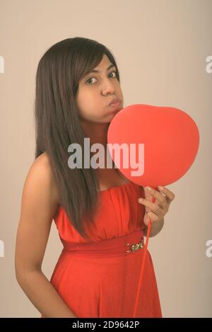 Studio shot der jungen Persischen Frau küssen, während rote herzförmige Ballons für den Valentinstag bereit, gegen grauer Hintergrund Stockfoto