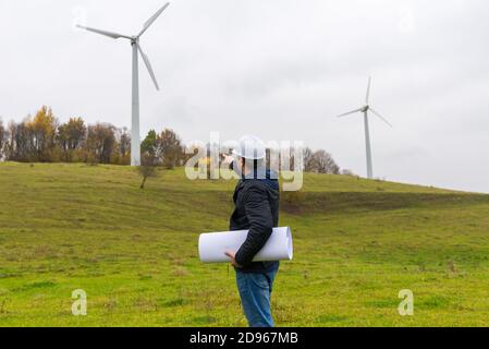 Rückansicht des Ingenieurs männlich zeigen auf die Windkraftanlagen Windmühlen, erneuerbare Energie und saubere Energie. Herbst wolkig Himmel grüne Wiese Stockfoto