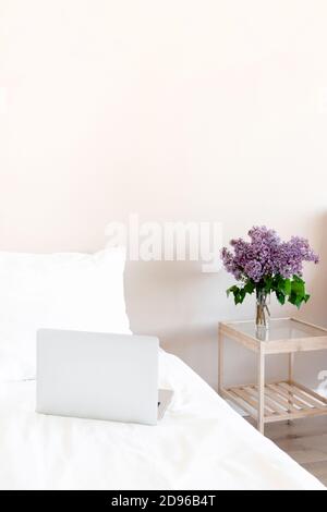 Helles Zimmer mit Laptop im Bett und Blumenstrauß Flieder auf einem Glastisch. Home-Arbeitsbereich. Stockfoto