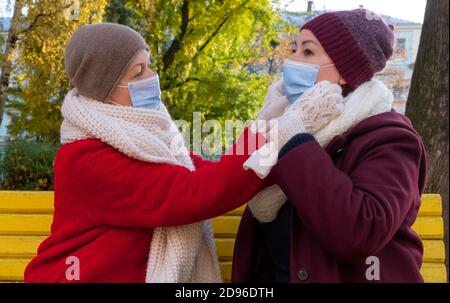 Glückliches Paar Senioren oder Frauen mittleren Alters tragen schützende medizinische Maske und sitzen auf einer Bank im Herbstpark. Stockfoto