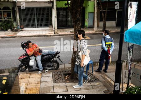 In einer typischen Nachbarschaftsszene fährt ein Mann mit Jeans die Straße hinunter, vorbei an einem Motorradtaxi und einem Wachmann. Stockfoto