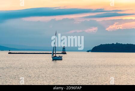 Sonnenuntergangsszene von Lake Champlain aus Burlington Vermont von einem Segelboot und Leuchtturm mit den Adirondack Mountains im Hintergrund. Stockfoto