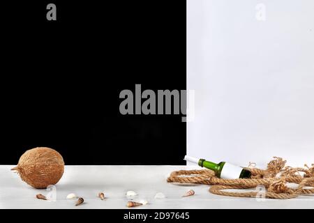 Reife Kokosnuss, Muschelschalen, Glasflasche mit Kopierraum und darin gerolltem Blatt Papier, dickes Seil auf Oberfläche vor schwarz-weißem Hintergrund Stockfoto