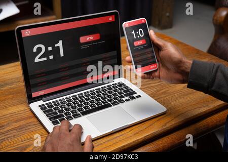 Nahaufnahme Laptop und Smartphone-Bildschirm mit Mobile App für Wetten und Gäste. Männliche Hände, die Geräte mit Matchergebnissen auf Bildschirmen halten. Glücksspiel, Wetten, Sport, Finanzen, moderne Technologien Konzept. Stockfoto