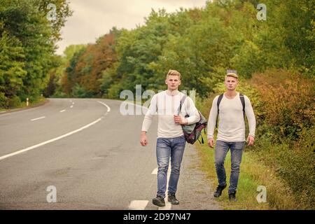 Firmenfreunde Reisende, die auf dem Naturhintergrund der Straße hitchwandern. Fahren Sie in der Nähe des Stadtrandes. Reisende auf dem Weg. Freunde, die hitchhitchhamps unterwegs Beginnen Sie mit dem Wandern ein tolles Abenteuer in Ihrem Leben. Stockfoto