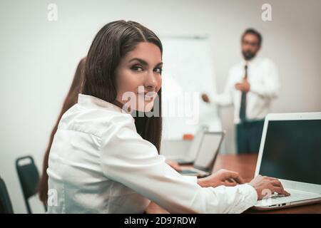 Hübsche Geschäftsfrau blickt zurück auf die Kamera, die am Laptop am Arbeitsplatz arbeitet. Männlicher Sprecher mit Präsentation des Projekts, der vor Weiß steht Stockfoto