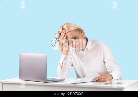 Erschöpfte reife Geschäftsfrau, die mit einem Laptop an ihrem Schreibtisch sitzt Und Dokumente auf blauem Studiohintergrund Stockfoto