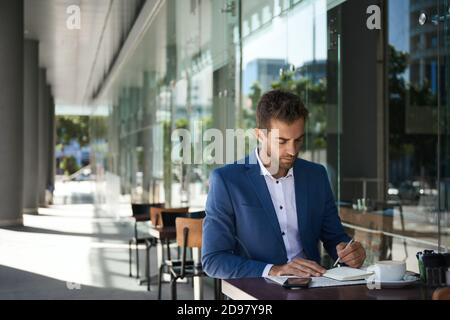 Geschäftsmann, der an einem Kaffeetisch sitzt und in einem Notizbuch schreibt Stockfoto