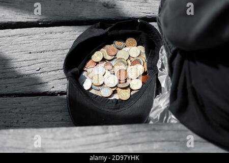 Ein Bettler sitzt neben einer Mütze mit Münzen und bittet um Almosen. Mütze mit ein paar Almosenmünzen Stockfoto