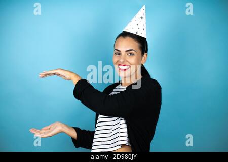 Junge schöne Frau trägt einen Geburtstagshut über insolated blauen Hintergrund gestikulieren mit Händen zeigt große und große Größe Zeichen, Maßnahme Symbol. Smil Stockfoto