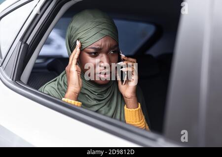 Gestresste schwarze muslimische Dame, die Auto auf dem Rücksitz reitet und spricht Auf dem Handy Stockfoto