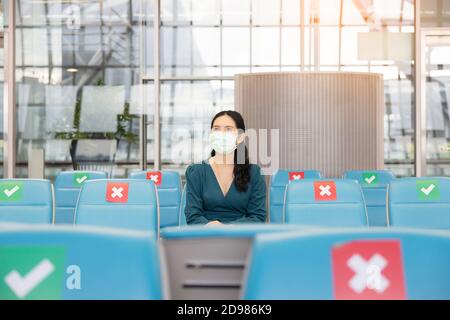 Asiatische Frau Tourist tragen Gesichtsmaske sitzen auf soziale Distanzierung Stuhl mit Gepäck wartet auf Flug am Flughafen Terminal während Coronavirus oder Kov Stockfoto