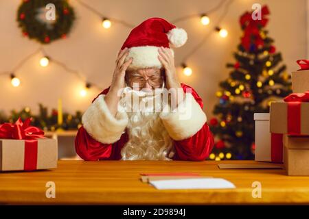 Trauriger alter bärtiger Weihnachtsmann, der an einem Tisch sitzt und seinen Kopf unter Kopfschmerzen hält. Stockfoto