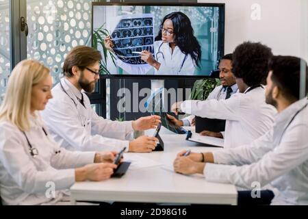 Team von multirassischen erfahrenen Ärzten oder Wissenschaftlern, mit Videokonferenz mit ihren jungen zuversichtlich afrikanischen Expertin und sprechen über die Stockfoto