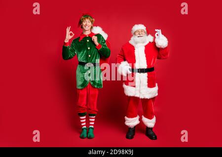 Foto in voller Länge von zwei Zeichen santa Elf halten Sack Zeige okey tragen x-Mas Kostüm Mütze Brille Schuhe isoliert rot Farbiger Hintergrund Stockfoto