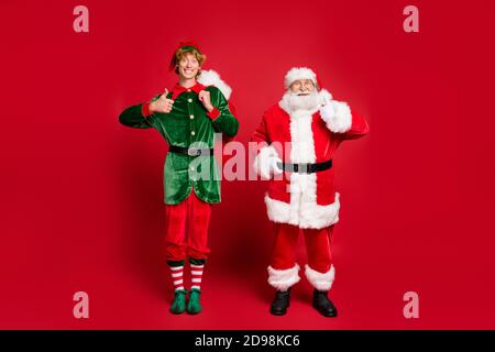 Foto in voller Länge von zwei Personen santa Elf halten groß Tasche erhöhen Daumen nach oben tragen x-Mas Kostüm Kappe Brille Stiefel Isolierter roter Hintergrund Stockfoto