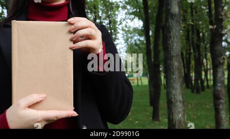 Nahaufnahme der Frau in schwarzem Wollmantel und Burgunder Pullover mit einem Buch mit leerem Einband im Park. Freier Platz für Ihr Lesekonzept Stockfoto
