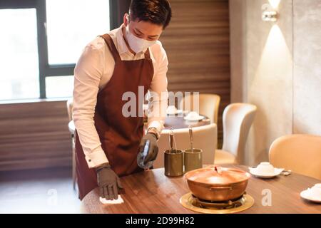 Der Kellner trägt eine Gesichtsmaske, während er im Restaurant desinfiziert Stockfoto