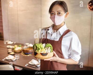 Junger Kellner trägt eine schützende Gesichtsmaske, während er Essen serviert Kunde im Restaurant Stockfoto