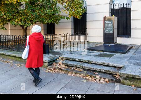 Eine Frau in einem roten Mantel geht am John F. Kennedy-Denkmal an der Marylebone Road vorbei, das aus Sicherheitsgründen aus seinem Sockel entfernt wurde, London, Großbritannien Stockfoto