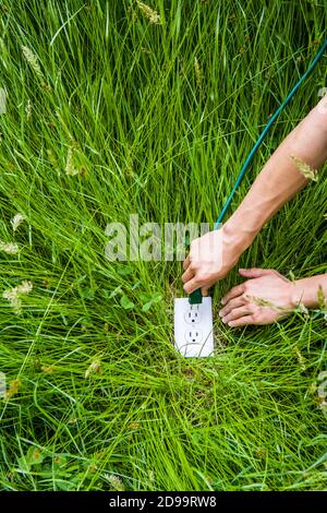 Eine konzeptuelle Aufnahme der grünen Energie. Ein mans Arme und Hände stecken ein Netzkabel an eine Steckdose im Boden umgeben von langen grünen Gras. Stockfoto