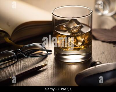Hintergrundbeleuchtetes Glas Whisky auf den Felsen auf einem Holztisch, mit Notizbuch, Stift und Lesebrille Stockfoto