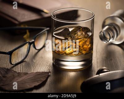 Hintergrundbeleuchtetes Glas Whisky auf den Felsen auf einem Holztisch, mit Notizbuch, Stift und Lesebrille Stockfoto